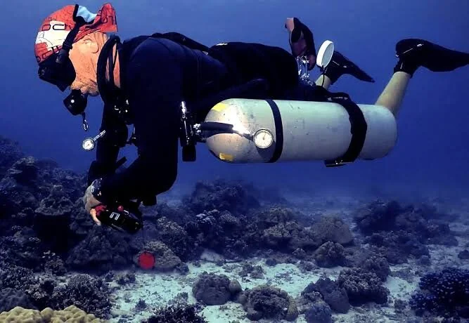 A diver wearing twin side-mounted cylinders reaches toward the reef below.