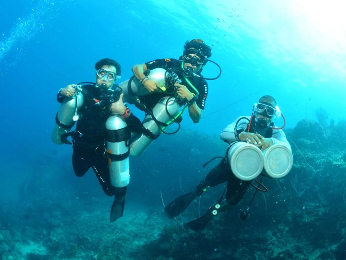 Three divers ascend together, one wearing twin cylinders mounted at the hips in sidemount style.