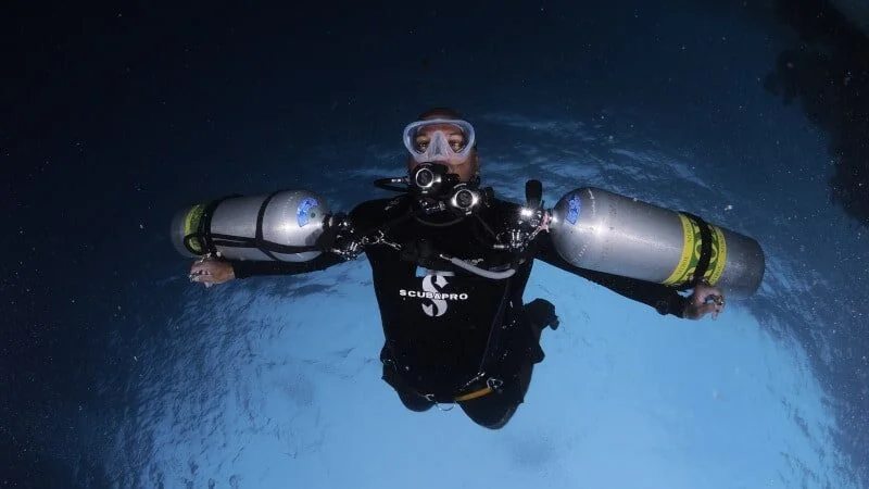 A diver floats facing the camera with a large cylinder strapped to each side of the body.