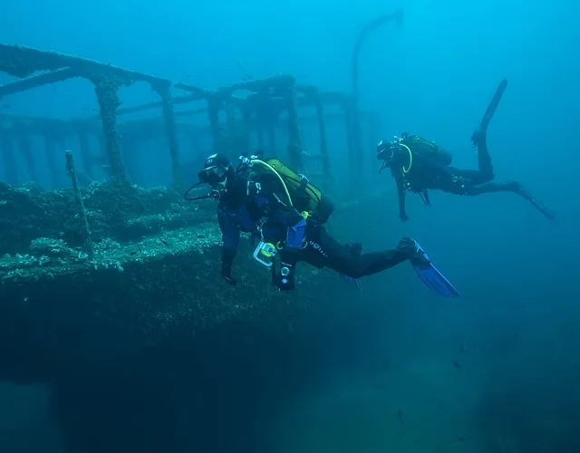 Two divers explore the coral-encrusted railings and superstructure of a large shipwreck.