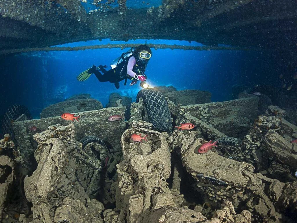 A diver shines a torch over debris and old tires scattered on a wreck's deck, with small red fish schooling nearby.