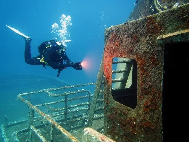 A diver aims a torch beam into a rusted doorway on a shipwreck's hull.