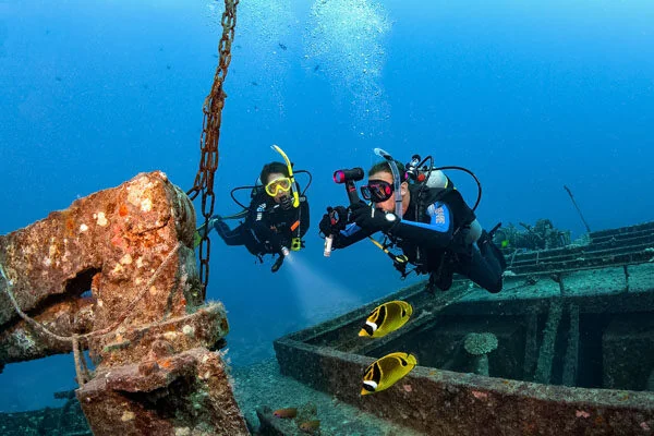 Two divers with torches examine a heavy rusted anchor chain hanging beside a shipwreck as fish swim past.