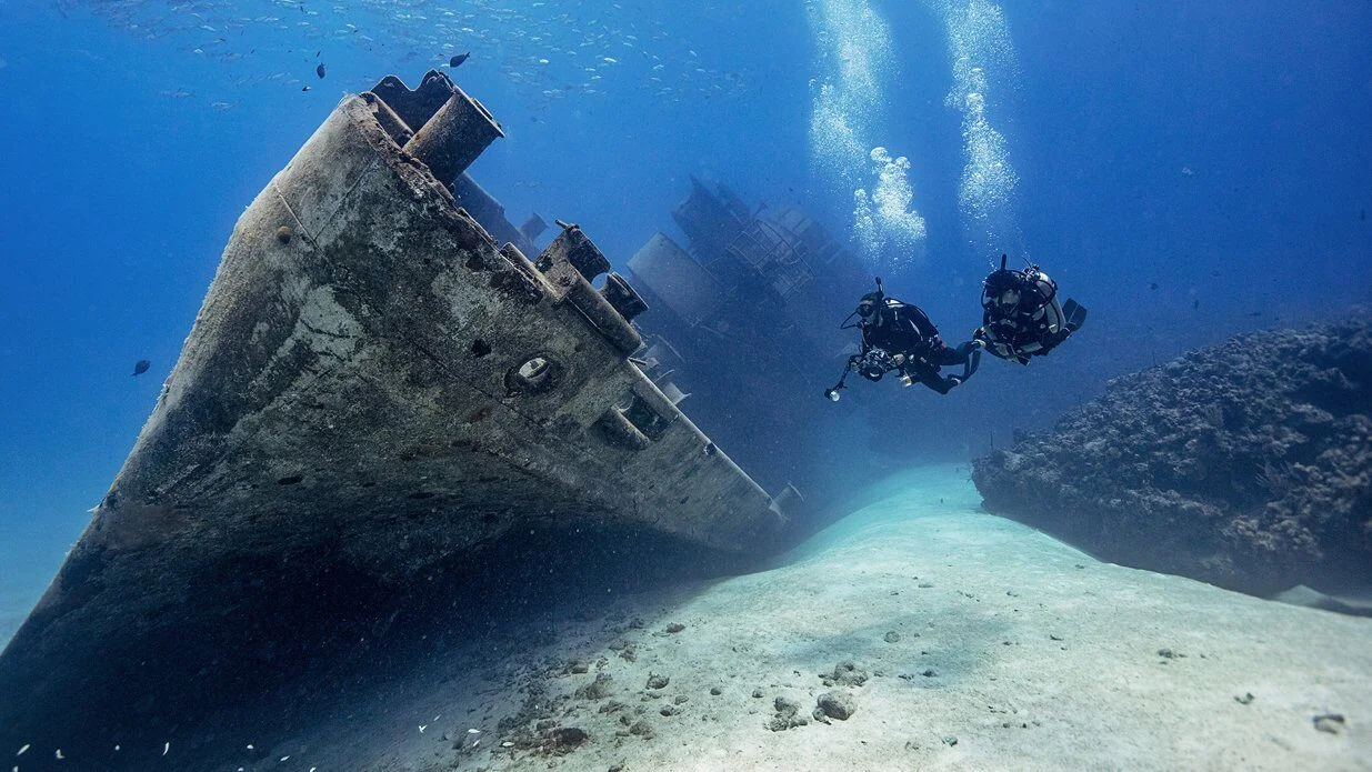 Two divers approach the towering upright bow of a large sunken ship surrounded by schooling fish.
