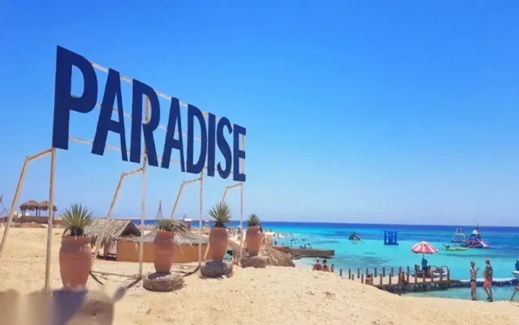A large blue PARADISE sign stands on the beach above straw cabanas and a crowded turquoise swimming area.