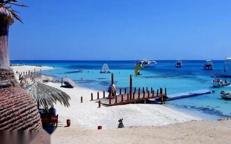 A wooden jetty leads toward moored boats and a shallow turquoise lagoon, seen under a straw umbrella awning.
