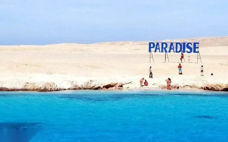 Tourists stand at the water's edge on a sandy dune below a PARADISE sign, with turquoise water in the foreground.