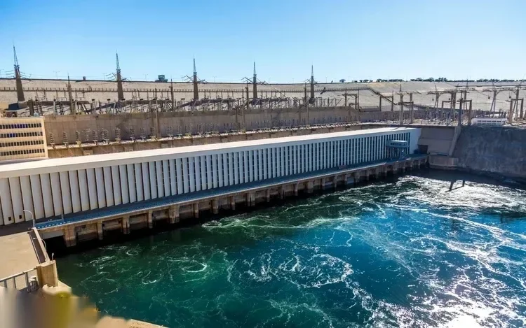 A wide view shows the Aswan High Dam's turbine hall and spillway with churning water below and power pylons along the top.