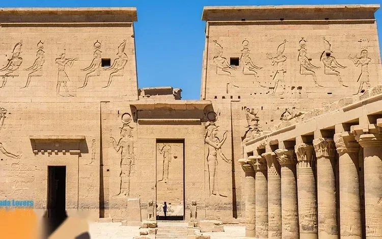 The carved facade of Philae Temple's first pylon depicts Egyptian gods, with a colonnade of columns beside it.
