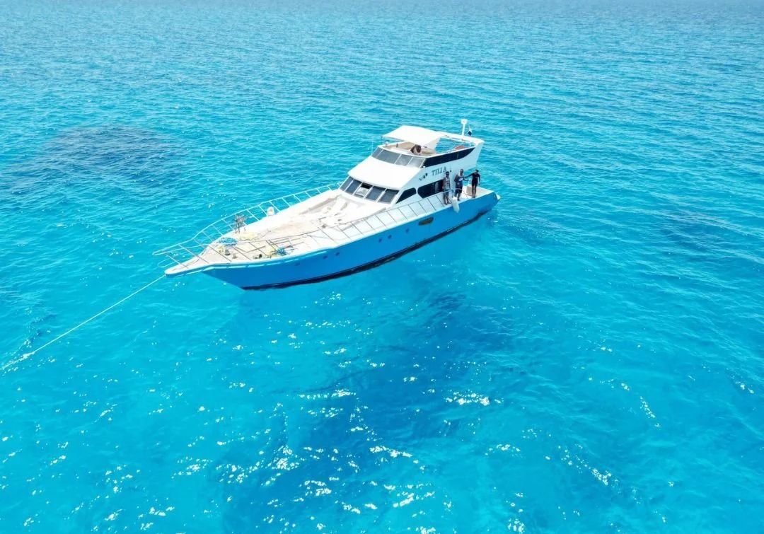 Aerial view of the white motor yacht anchored in bright turquoise water with two crew members standing on deck.