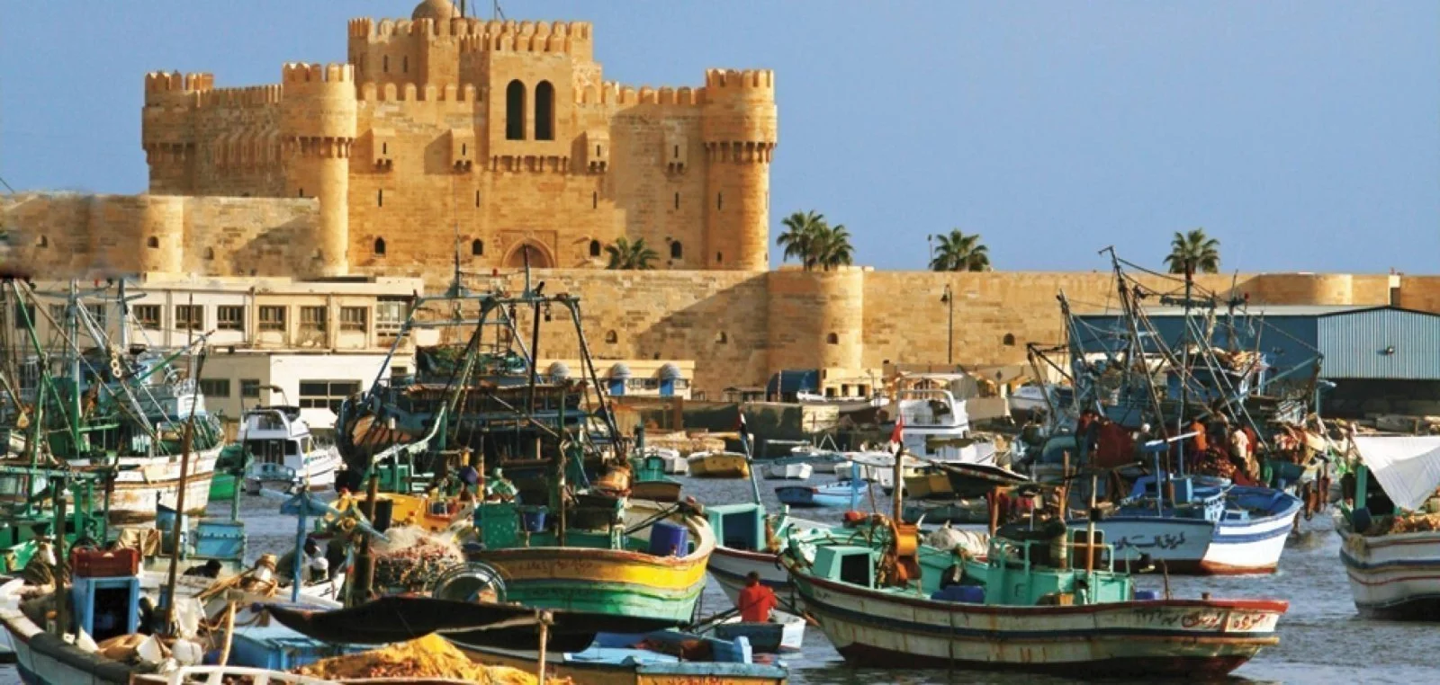 Qaitbay Citadel's sandstone fortress walls rising behind a crowded harbor of colorful wooden fishing boats