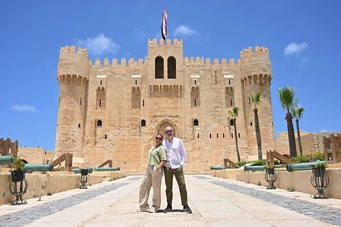 Couple posing arm in arm on the stone causeway in front of Qaitbay Citadel with the Egyptian flag flying above its towers
