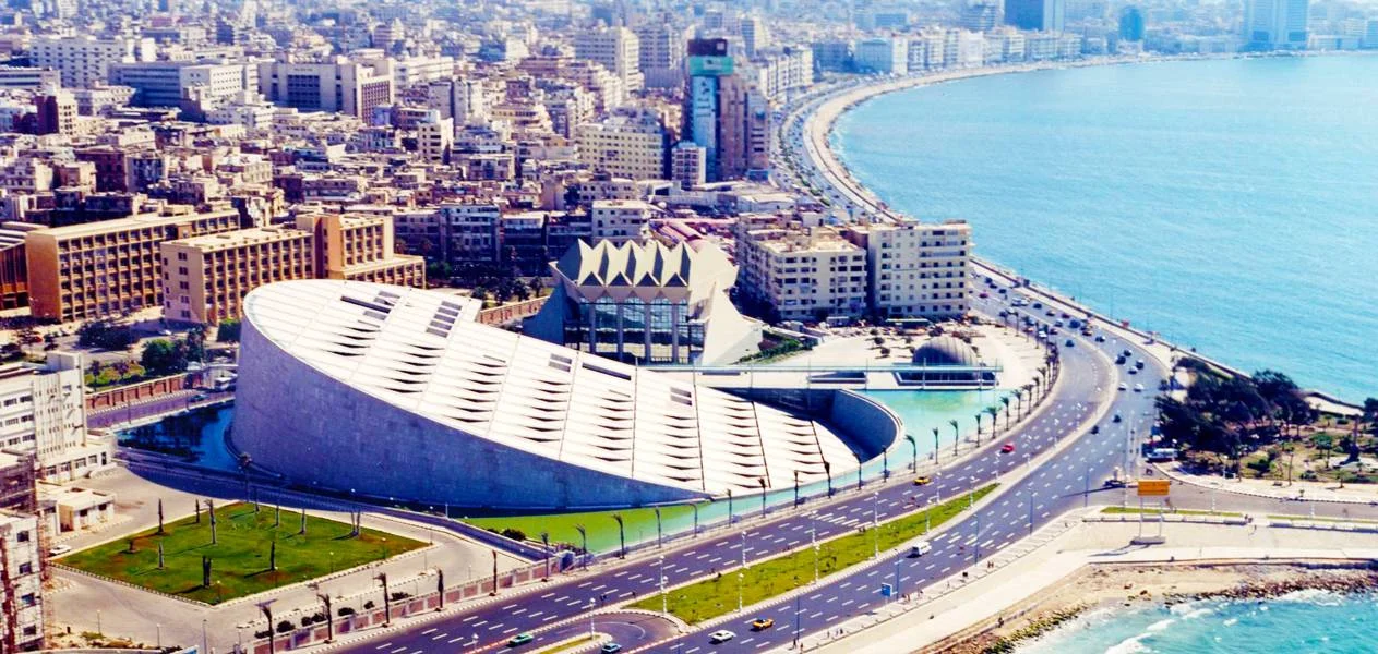 Aerial view of Alexandria's corniche showing the fan-shaped Bibliotheca Alexandrina library building beside the curving Mediterranean coastline and city skyline