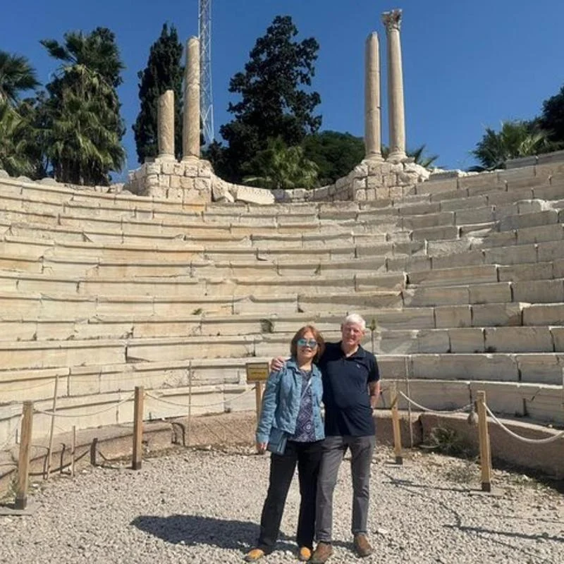 Couple standing among the weathered limestone tiers of the Roman Amphitheatre (Kom el-Dikka) with ancient columns and palm trees behind them