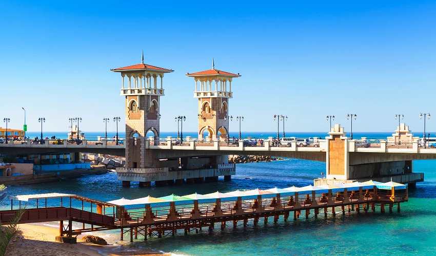 Stanley Bridge in Alexandria with its twin Ottoman-style towers spanning turquoise Mediterranean water, beach umbrellas and a wooden pier in the foreground