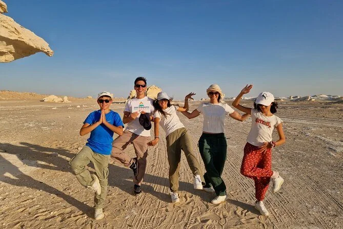 Five friends pose playfully with raised arms and yoga-style poses in the White Desert's sand among chalky rock formations.