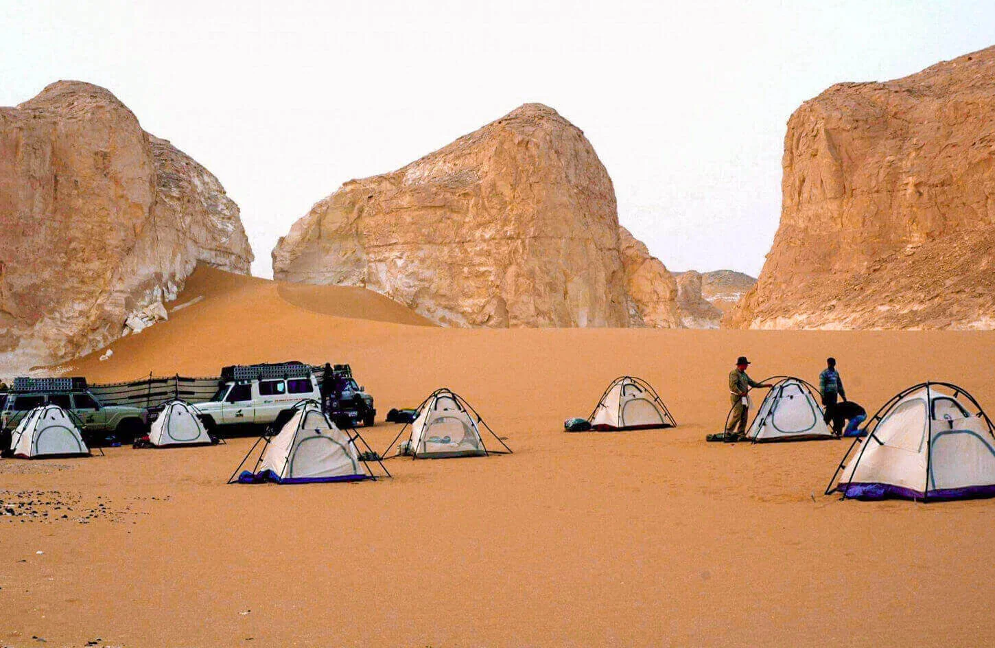 Several dome tents are pitched on sand among towering white chalk rock formations for an overnight desert camp.