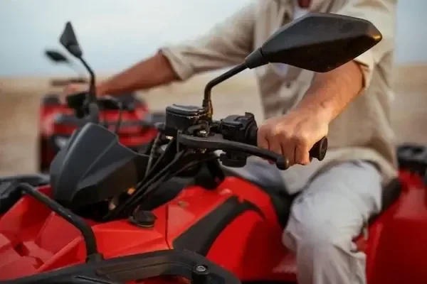 Close-up of a man's hands gripping the handlebars of a red quad bike, another rider visible behind.