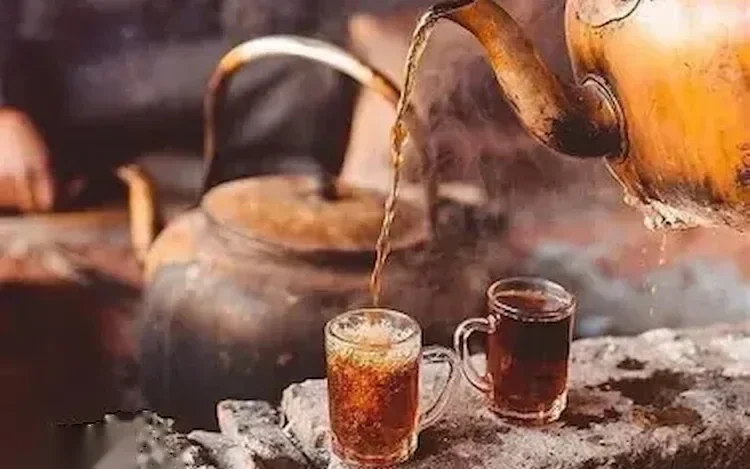 Traditional metal kettle pouring hot tea into two glass cups over hot coals.