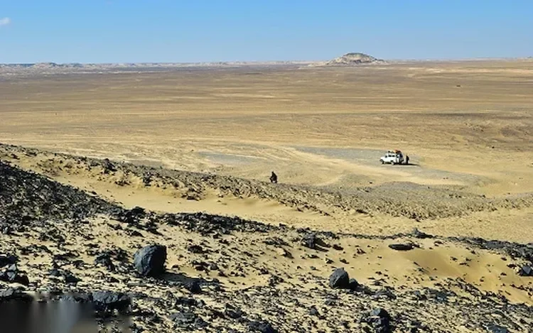 Wide view of a flat rocky desert plain with a parked 4x4 vehicle and two small figures in the distance.