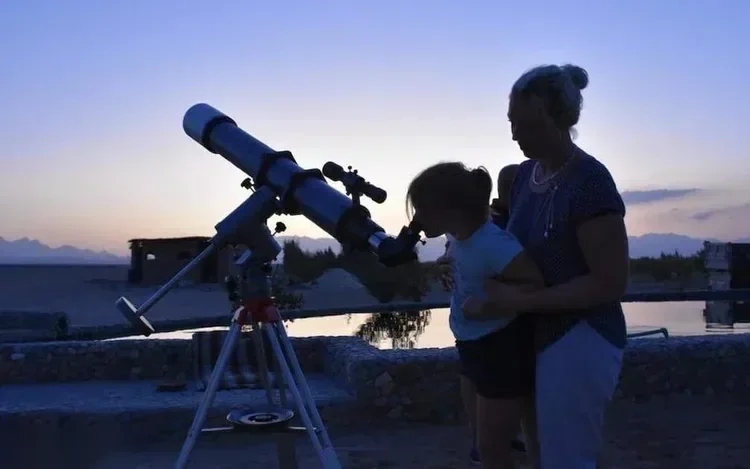 Silhouette of a woman and child looking through a telescope set up outdoors at dusk.