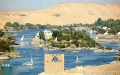Wide view of the Nile at Aswan with feluccas sailing past granite boulders and a domed riverside building.