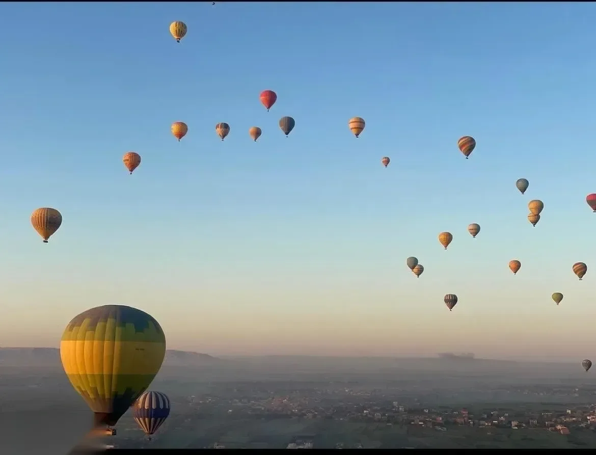 Dozens of hot air balloons scattered across the sky over the Luxor valley at sunrise, seen from inside another balloon's basket.