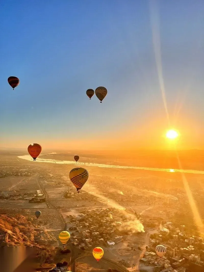 Hot air balloons floating above the Nile valley at sunrise with the sun low over the horizon and the river below.
