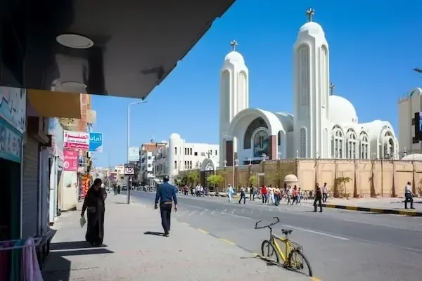 A Hurghada street with a Coptic church's twin white bell towers topped with crosses, pedestrians and a bicycle nearby.