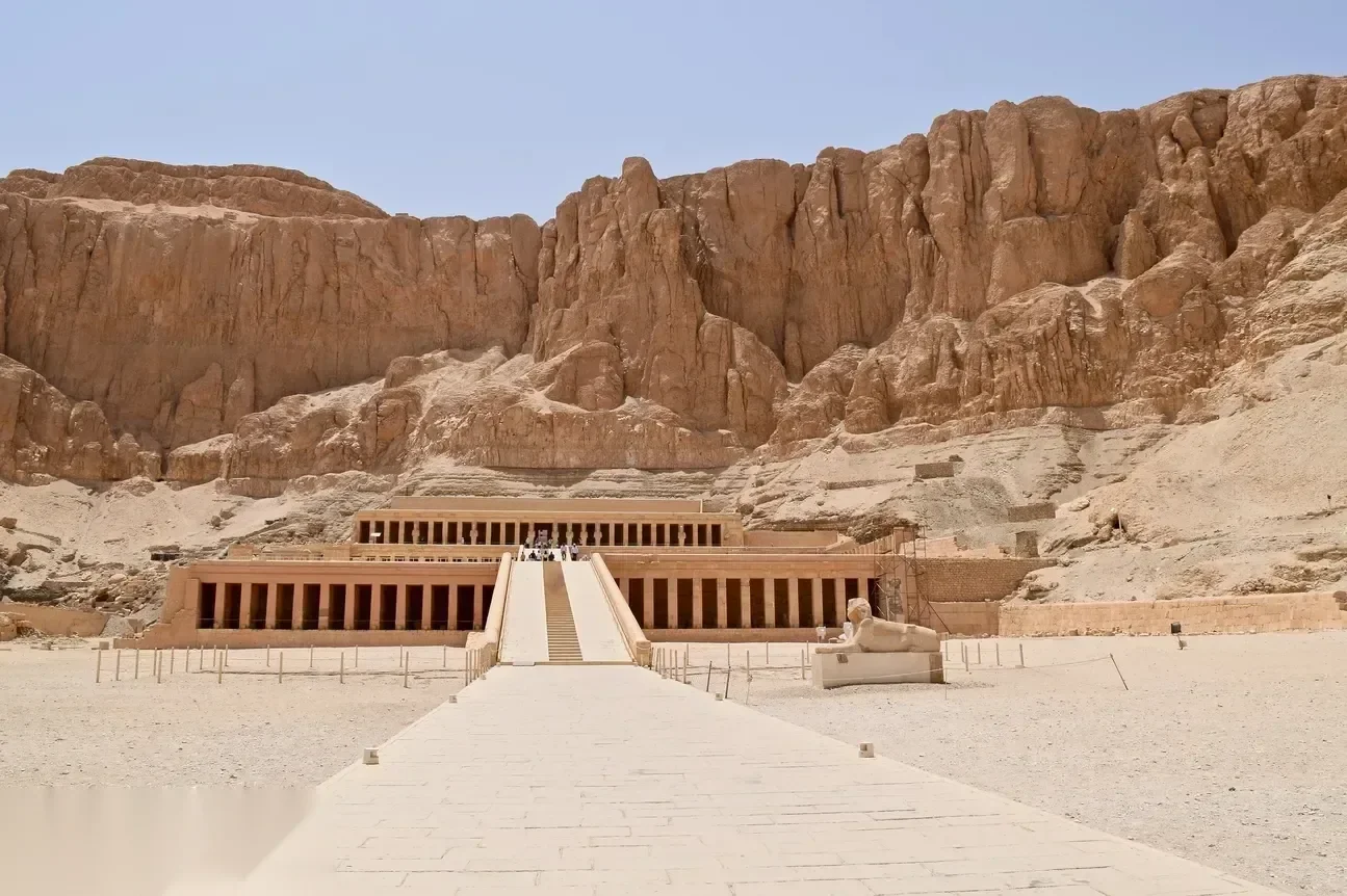 A long paved causeway leads up to the three-tiered Temple of Hatshepsut, built into sheer cliffs at Deir el-Bahari, with a small sphinx statue beside the ramp.