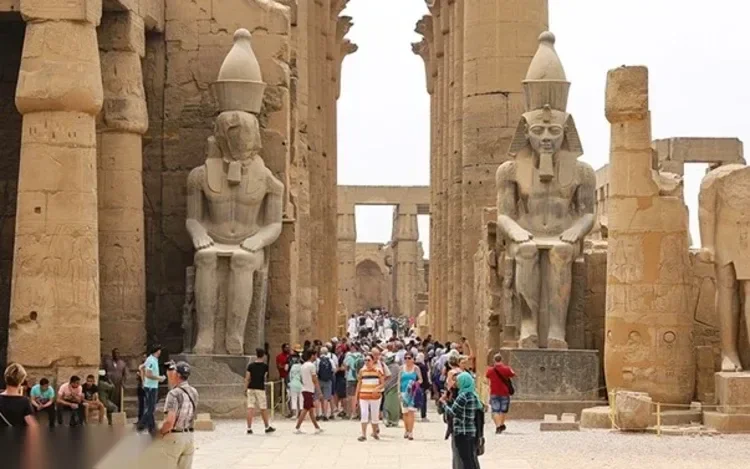Colossal seated pharaoh statues flank a temple corridor crowded with tourists in white and blue clothing, viewed from nearly the same angle as another photo in this set.