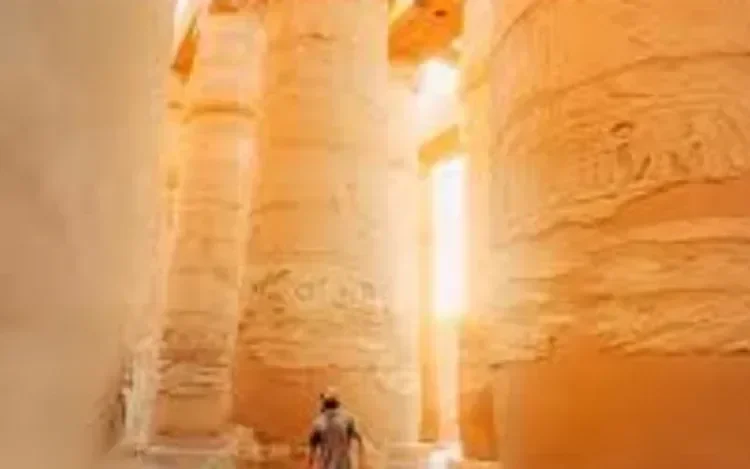 Sunlight streams between towering stone columns inside a temple hall, silhouetting a single visitor walking through the golden haze.