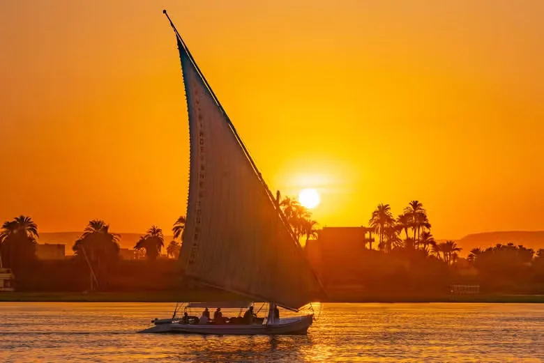 A traditional felucca sailboat with a tall white sail crosses the Nile in Luxor as the sun sets behind silhouetted palm trees.