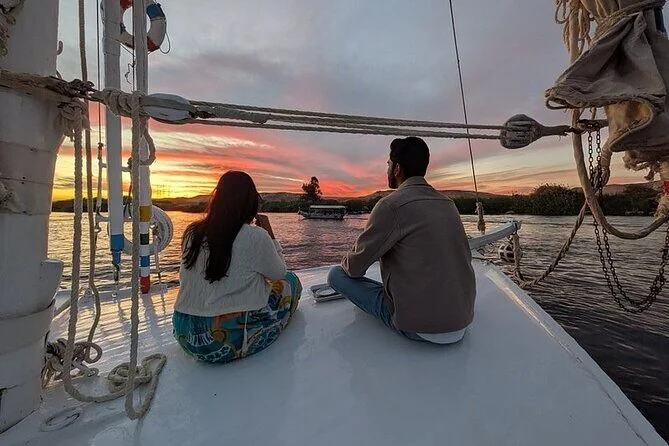 A couple sits at the bow of a felucca watching a pink and orange sunset over the Nile in Luxor.