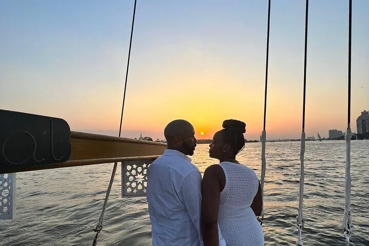 A couple stands close together at the front of a felucca, silhouetted against the setting sun over the Nile.