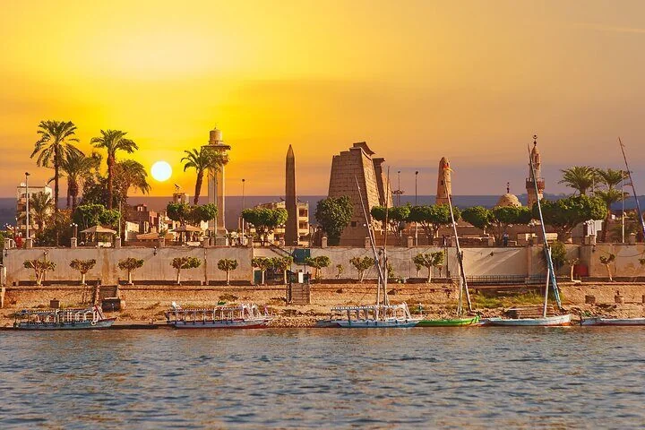 Feluccas are moored along the Luxor riverbank at sunset, with the obelisk and pylon of Luxor Temple visible above the palm trees.