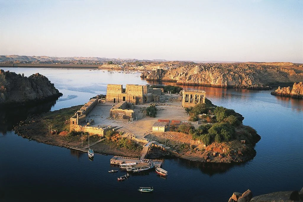 Aerial view of Philae Temple's island complex surrounded by the waters of Lake Nasser, with small tour boats docked at its shore.