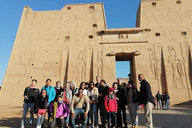 A large group of tourists poses for a photo in front of the massive carved pylon entrance of Philae Temple.