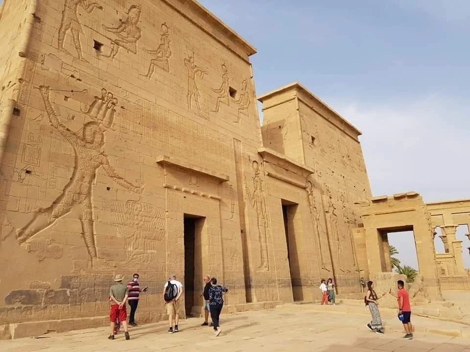 Visitors walk past the carved outer wall of Philae Temple, decorated with reliefs of Egyptian gods and a pharaoh, with a further temple gateway visible beyond.