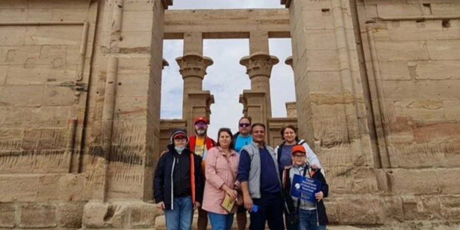A family group poses together in the entrance gateway of Philae Temple, with columns and pylon towers framing them.