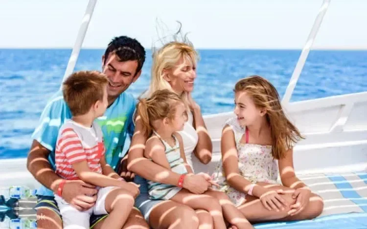 A family of two adults and three children sit together laughing on the deck of a sailing yacht at sea.