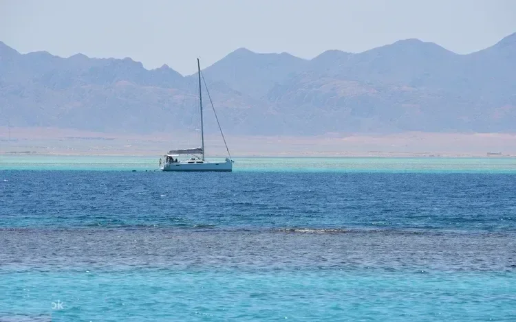 A white sailboat is anchored near a reef with desert mountains along the coastline in the background.