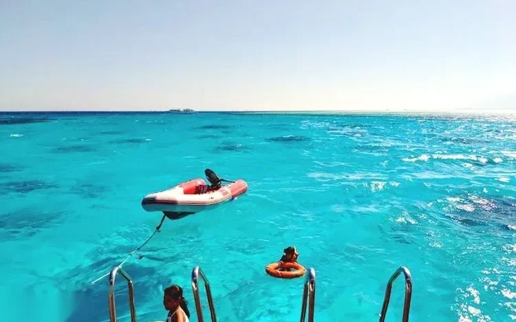 View from a boat's swim ladder toward a small inflatable tender boat and a red buoy floating on bright turquoise water.