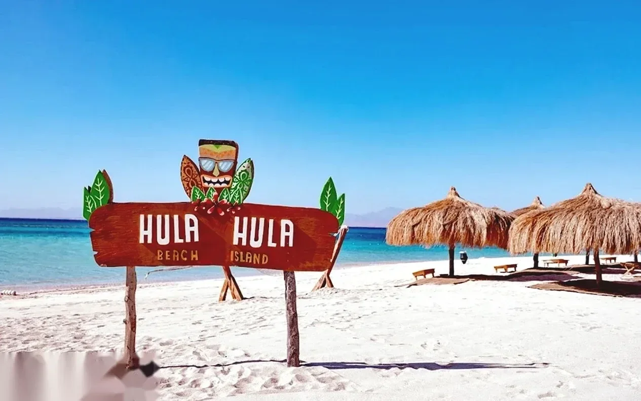 A wooden Hula Hula Beach Island sign on a white sand beach, with straw-thatched umbrellas and turquoise water beyond.