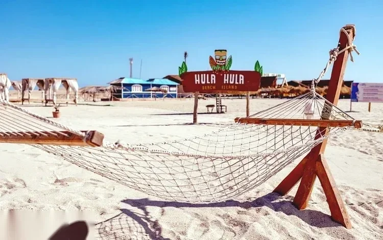 A rope hammock strung between wooden posts on the sand, with the Hula Hula Beach Island sign and thatched huts in the background.