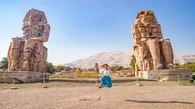 Woman in a blue skirt jumping for a photo between the two Colossi of Memnon statues under a clear sky.