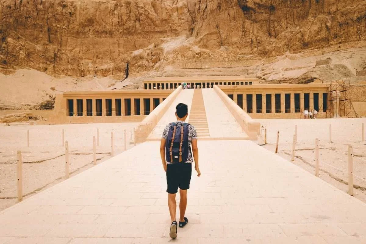 Man with a backpack walking along the ramp toward the Temple of Hatshepsut set into the cliffs.