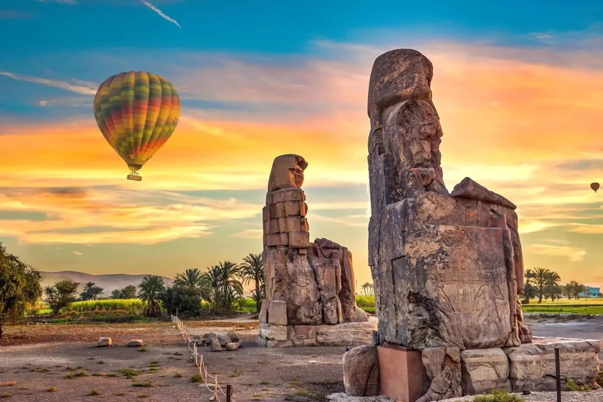 The two Colossi of Memnon statues at sunset with a hot air balloon floating in the colorful sky behind them.