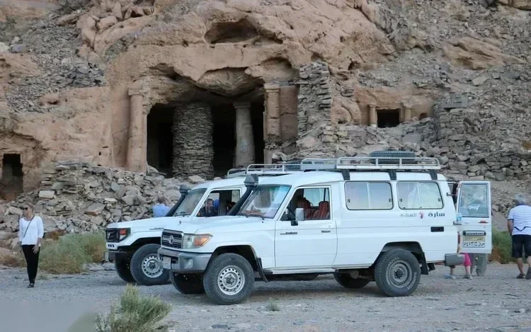 Two white 4x4 vehicles parked in front of ancient rock-cut ruins with stone columns.