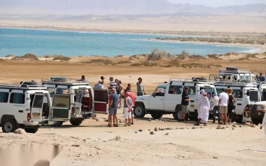 Convoy of 4x4 vehicles parked beside a turquoise lagoon with tourists and a Bedouin guide gathered nearby.
